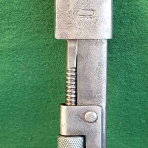 May include: A silver adjustable wrench with a knurled handle and a threaded adjustment screw. The wrench has a  circular opening on the top and a small, circular opening on the side.