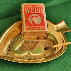 Puede incluir: Un cenicero de latón dorado con forma de hoja, con una caja de cerillas "CAPTAIN WEBB". La caja es roja y beige, con un retrato de un hombre. Una cerilla quemada descansa en el cenicero.
