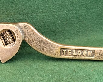 Puede incluir: Una llave inglesa de latón vintage con la palabra "TELCON" estampada en el mango. La llave ajustable tiene un mango curvo y una mordaza ajustable. La llave está sobre una superficie verde.