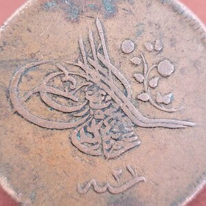 May include: Close-up of a copper coin with Arabic script and a floral design. The coin is worn and has a green patina.