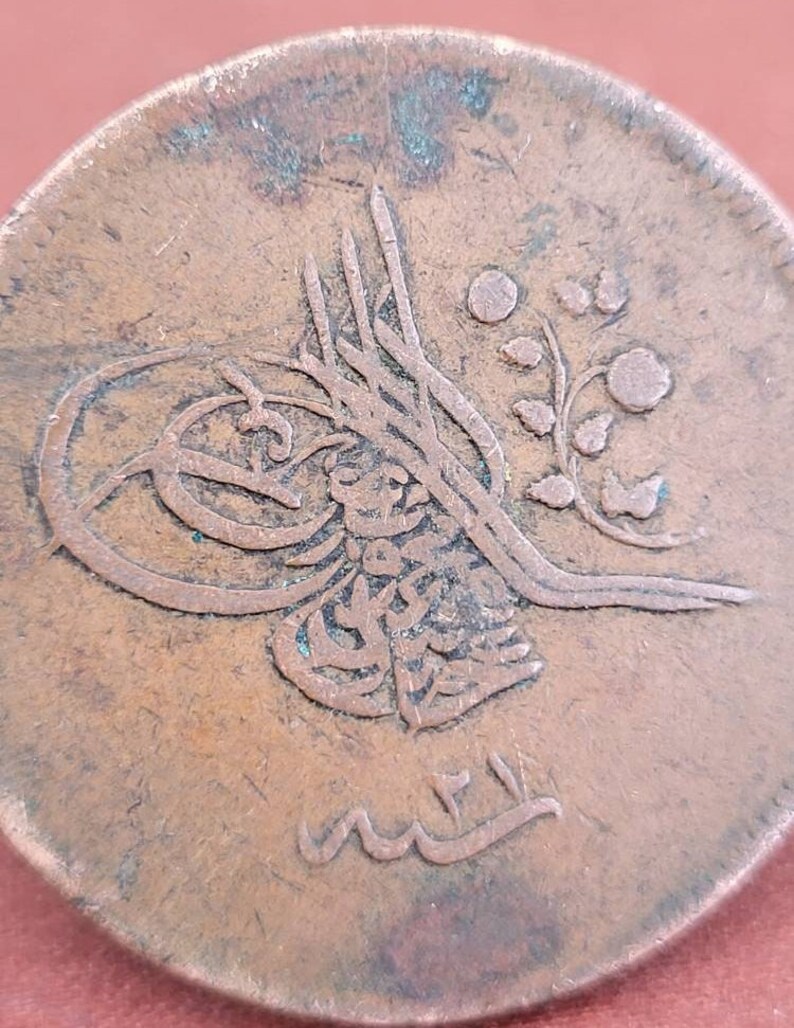 May include: Close-up of a copper coin with Arabic script and a floral design. The coin is worn and has a green patina.