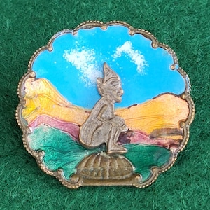 May include: A vintage, ornate pin featuring a seated figure with a pointed hat against a backdrop of a blue sky with clouds and a colorful landscape. The pin has a gold-toned metal frame with an intricate border and is set on a green background.