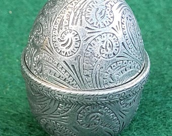 Puede incluir: Un joyero de metal plateado con forma de huevo que presenta un intrincado patrón de cachemira en relieve en toda la superficie. El recipiente consta de dos mitades que encajan entre sí y descansa sobre una pequeña base central.