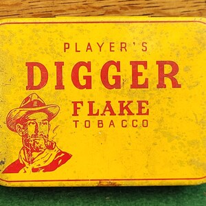 May include: A rectangular yellow tin with rounded corners. The tin features the words "PLAYER'S DIGGER FLAKE TOBACCO" in red letters. An illustration of a man in a hat smoking a pipe is on the left side of the tin.
