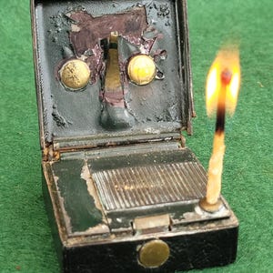 Puede incluir: Un antiguo cerillero abierto, que revela la superficie de fricción y dos detalles de latón. Un cerilla encendida con una llama naranja brillante se coloca junto al cerillero. El cerillero es negro y verde con una tapa con bisagras.