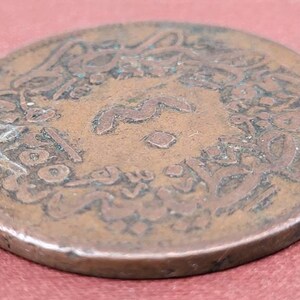 May include: A copper coin with Arabic script around the edge. The coin is worn and has a greenish patina.