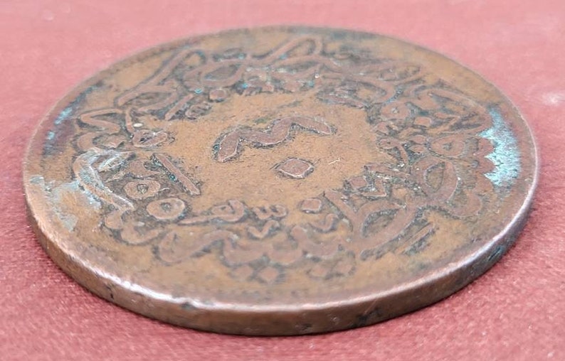 May include: A copper coin with Arabic script around the edge. The coin is worn and has a greenish patina.