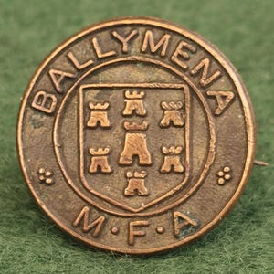 May include: A circular bronze-toned metal button featuring the embossed text BALLYMENA at the top and M.F.A at the bottom. The center displays a shield emblem containing five small castle tower icons arranged in a cross pattern.
