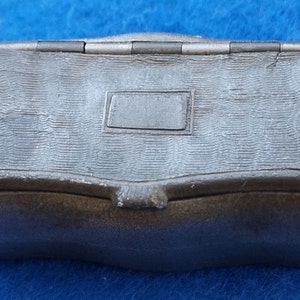 Op de afbeelding: Een klein, grijs metalen doosje met een scharnierend deksel. Het doosje heeft een getextureerd oppervlak en een rechthoekige, lege plaquette aan de bovenkant.