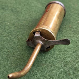 May include: A vintage brass oiler featuring a cylindrical body with a knurled cap and a curved, tapered spout. The mechanism includes a metal lever attached to the base of the spout, designed for manual operation to dispense oil from the container.