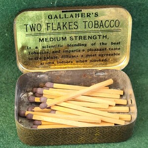 Puede incluir: Una lata de metal dorada para tabaco con el texto "GALLAHER'S TWO FLAKES TOBACCO MEDIUM STRENGTH" en la tapa. La lata está abierta y llena de cerillas de madera.