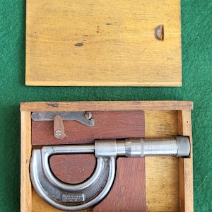 May include: A silver micrometer in a wooden box with a red felt lining. The micrometer has a scale marked with numbers and a small handle. The box has a hinged lid.
