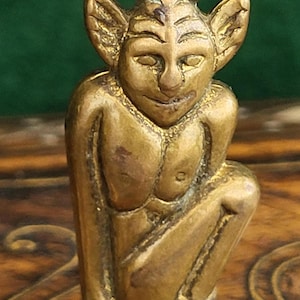 Könnte beinhalten: Kleine, messingfarbene Figur eines hockenden Goblins oder Kobolds. Die Figur hat große, spitze Ohren, ein lächelndes Gesicht und kniet. Sie hat oben eine Schlaufe zum Aufhängen. Die Figur ist detailliert und scheint aus der Vintage-Zeit zu stammen.