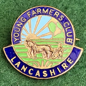 Puede incluir: Un pin vintage del Young Farmers Club Lancashire. El pin presenta un borde dorado con un fondo de esmalte azul. El centro representa una escena de granja con un sol naciente, árboles y un granjero arando un campo. El texto "Young Farmers Club" y "Lancashire" es azul.
