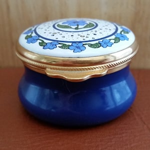 Puede incluir: Una caja de porcelana azul y blanca con un diseño floral. La tapa está decorada con flores de nomeolvides y un borde dorado. La caja tiene una bisagra dorada y un borde festoneado.