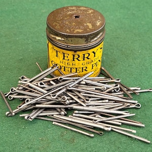 May include: A tin of Terry's High Grade Cotter Pins. The tin is yellow with black lettering and is sitting on a green surface. The tin is surrounded by a pile of silver cotter pins.