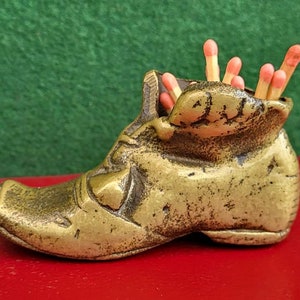 Puede incluir: Un porta-cerillas de metal de color dorado en forma de bota con un acabado desgastado. El porta-cerillas está lleno de cerillas con puntas rosas.