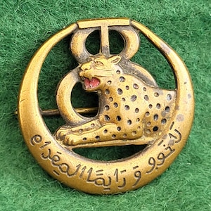 以下が含まれることがあります： 詳細なヒョウのデザインが施されたヴィンテージ真鍮ピン。赤いアクセントとアラビア語のスクリプトが特徴です。ピンは円形のフレーム内に配置され、上部に様式化されたエンブレムが付いています。背景は緑色です。