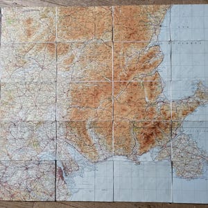 May include: A folded vintage map, divided into rectangular panels, displaying a detailed topographical view. The map uses a colour scheme of tan, brown, and blue, with intricate lines indicating roads and geographical features.