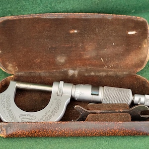 May include: A silver-coloured micrometer with a brown leather case. The micrometer has a dial with markings in inches and a scale that reads up to 20. The case has the text "Moore & Wright Sheffield England" on it.