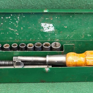 Puede incluir: Una caja de herramientas de metal verde con un destornillador de mango de madera y un juego de vasos de metal. La caja de herramientas está abierta y las herramientas son visibles en el interior.