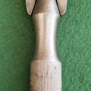 Może przedstawiać: Srebrne metalowe narzędzie z radełkowanym uchwytem i zakrzywioną, otwartą głowicą. Narzędzie jest przeznaczone do określonego celu, prawdopodobnie związanego z kształtowaniem lub cięciem materiałów. Narzędzie jest pokazane na zielonym tle.