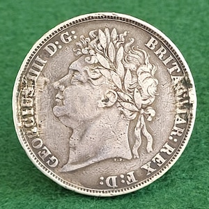 May include: A silver coin depicting a profile of a man with a laurel wreath, encircled by inscriptions. The coin shows signs of wear, indicating its age. The coin is set against a green backdrop.