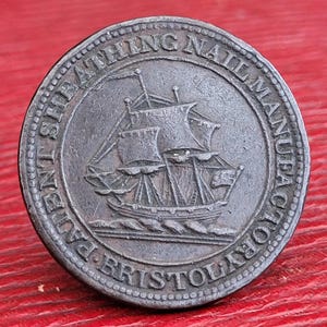 May include: A circular, bronze-colored metal token with a ship design and the text "SHEATHING NAIL MANUFACTORY BRISTOL" around the edge.