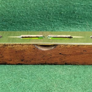 Puede incluir: Un nivel de madera vintage con una parte superior de latón y dos niveles de burbuja. El nivel mide aproximadamente 30 cm de largo.