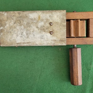 Puede incluir: Un juguete de madera con un panel de metal y una manivela. El juguete tiene un marco de madera y un panel de metal con dos tornillos. La manivela es de madera y tiene una forma hexagonal.