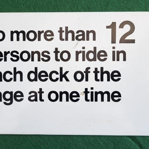 Può includere: Un cartello rettangolare bianco con testo nero: "Non più di 12 persone per gabbia alla volta". Il cartello ha quattro piccoli fori, uno in ogni angolo. Lo sfondo è verde.