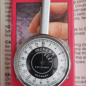 May include: A map measurer with a silver handle and a circular dial. The dial displays measurements in inches and feet, with a range from 0 to 91 cm. The text "COPYRIGHT GERMANY" is visible on the dial. The device is set against a red background.