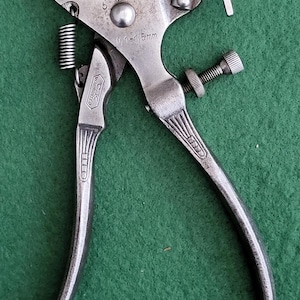 May include: A vintage metal tool with a spring, screw, and curved handles. The tool has the text "0.1-1.5mm" and "C.K. WARRANTED" stamped on it. The tool is silver and sits on a green surface.