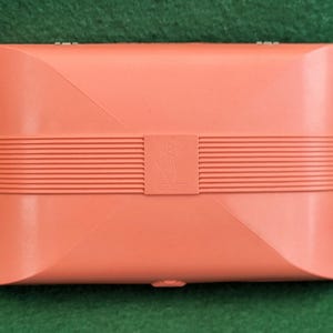 May include: A coral-coloured rectangular box with a ridged band across the centre and a small emblem. The box has a curved design on the top and bottom. The background is a solid green colour.