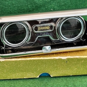 May include: Vintage silver-toned opera glasses with the word "DIAMOND" on the front. The glasses are open, revealing the lenses, and are resting on a gold-colored box. The box has a blue accent on the bottom.
