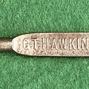 May include: A vintage metal button hook with a loop at one end and a rounded tip at the other. The hook has the text "G.T. HAWKINS NORTHAMPTON" engraved on the handle. The button hook is silver in color.