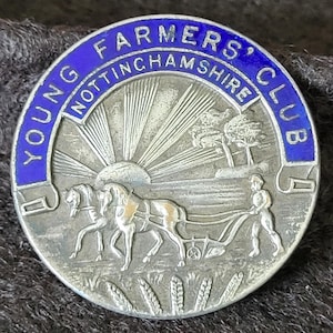 Puede incluir: Una insignia plateada del Young Farmers' Club con un borde de esmalte azul. La insignia presenta una escena de un agricultor arando un campo con dos caballos, un sol naciente y el texto "YOUNG FARMERS' CLUB NOTTINGHAMSHIRE".