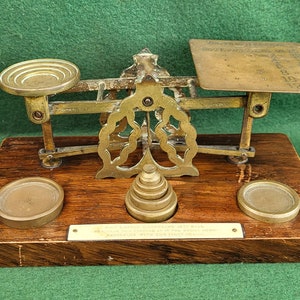 May include: Antique brass balance scale with a wooden base and a brass plate with markings for weights. The scale has a decorative design and a small plaque that reads "WE DO NOT RECOMMEND TESTING THIS WITH PRECIOUS METAL FOR YOUR FIRST TRY."
