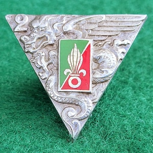 May include: Silver-toned triangular pin with a central emblem of green, red, and white. The pin features a dragon and wing design, with the number "2" at the top left. The background is a green surface.