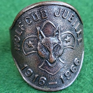 Könnte beinhalten: Ein Vintage-Ring in Bronzetönen mit den Worten "WOLF CUB JUBILEE" und den Jahren "1916-1966" geprägt. Der Ring zeigt einen detaillierten Wolfskopf in der Mitte, mit Sternen auf beiden Seiten. Der Ring ist vor einem grünen Hintergrund platziert.