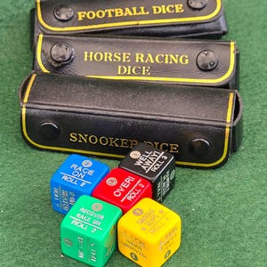 Puede incluir: Tres estuches rectangulares negros con cierres a presión, etiquetados como Football Dice, Horse Racing Dice y Snooker Dice. Cinco dados multicolores en azul, rojo, negro, verde y amarillo dispuestos delante, cada uno con instrucciones.