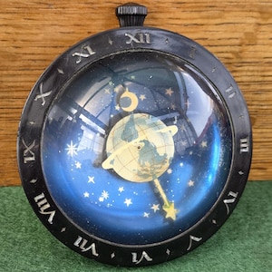 Könnte beinhalten: Eine runde Uhr mit schwarzem Gehäuse und einem dunkelblauen Zifferblatt mit Himmelsmotiv. Das Zifferblatt zeigt eine zentrale Globus-Illustration, umgeben von Sternen und einer Mondsichel. Ein schwarzer Außenring zeigt silberne römische Ziffern.