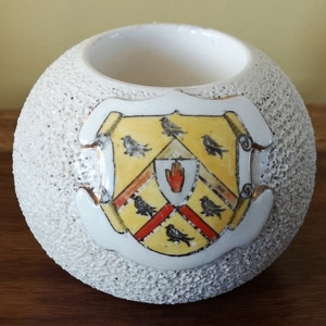 Puede incluir: Portavelas de cerámica blanca con superficie texturizada. El portavelas presenta un escudo con borde dorado, fondo amarillo, rayas rojas y blancas y pájaros negros.