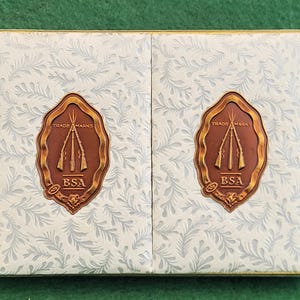 Puede incluir: Caja rectangular con un estampado floral blanco y gris. La caja tiene dos paneles, cada uno con un emblema ovalado marrón con tres rifles y el texto "TRADE MARKS" y "BSA". La caja tiene un borde dorado.