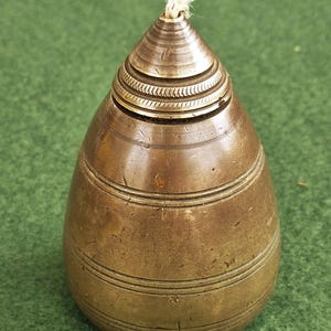 Può includere: Un contenitore vintage in ottone con una parte superiore conica e una base arrotondata. Il contenitore presenta fasce orizzontali e un design strutturato vicino alla parte superiore. Un filo bianco sporge dalla parte superiore. Il contenitore è su una superficie verde.