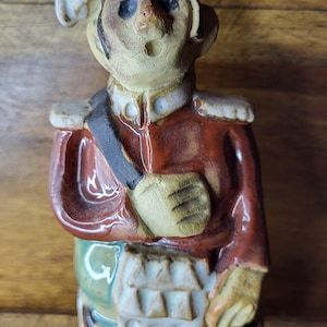 Könnte beinhalten: Keramikfigur eines Kochs in roter Jacke, schwarzer Hose und weißer Mütze. Die Figur hat ein detailliertes Gesicht und hält eine weiße Schürze. Die Figur steht auf einer Holzoberfläche.