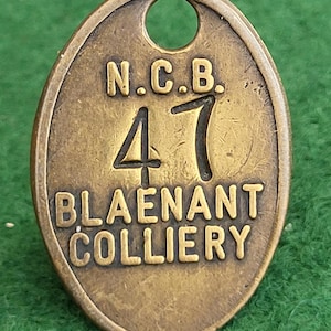 May include: Oval-shaped brass tag with the text "N.C.B. 47 BLAENANT COLLIERY" embossed on it. The tag has a hole at the top for attachment. The tag is set against a green background.