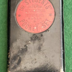 May include: A vintage black metal pocket set case with a red circular label. The label reads "The Students Criterion Mathematical Instruments Pocket Set" with text and star accents. The case shows signs of wear.