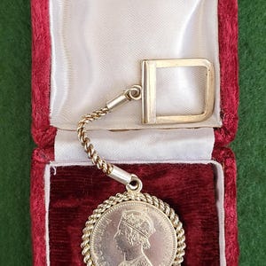 May include: A silver coin keychain with a portrait of Queen Victoria on the front, in a red velvet box with a white satin lining.