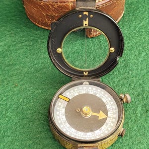 May include: An open, antique compass with a black and brass finish, featuring a circular dial with directional markings. The compass is set against a green background, with a leather case visible in the background.
